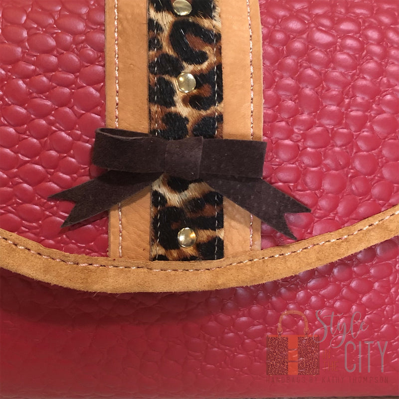 Close up of front flap details of red leather handbag with leopard trim and leather bow & gold nailheads.