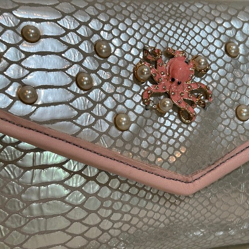 Close up view of pearl studs and pink octopus accent piece on silver snake print leather clutch. 